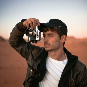 Focused young male traveler in stylish outfit and cap taking photo on vintage camera during trip in desert valley during sunset