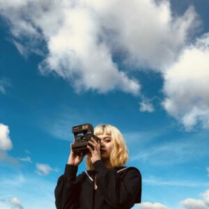 Low angle of young blond lady in trendy black outfit standing under blue cloudy sky with vintage photo camera in hands in daylight