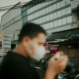 Side view of blurred unrecognizable male in medical mask with photo camera standing on street in local district during pandemic